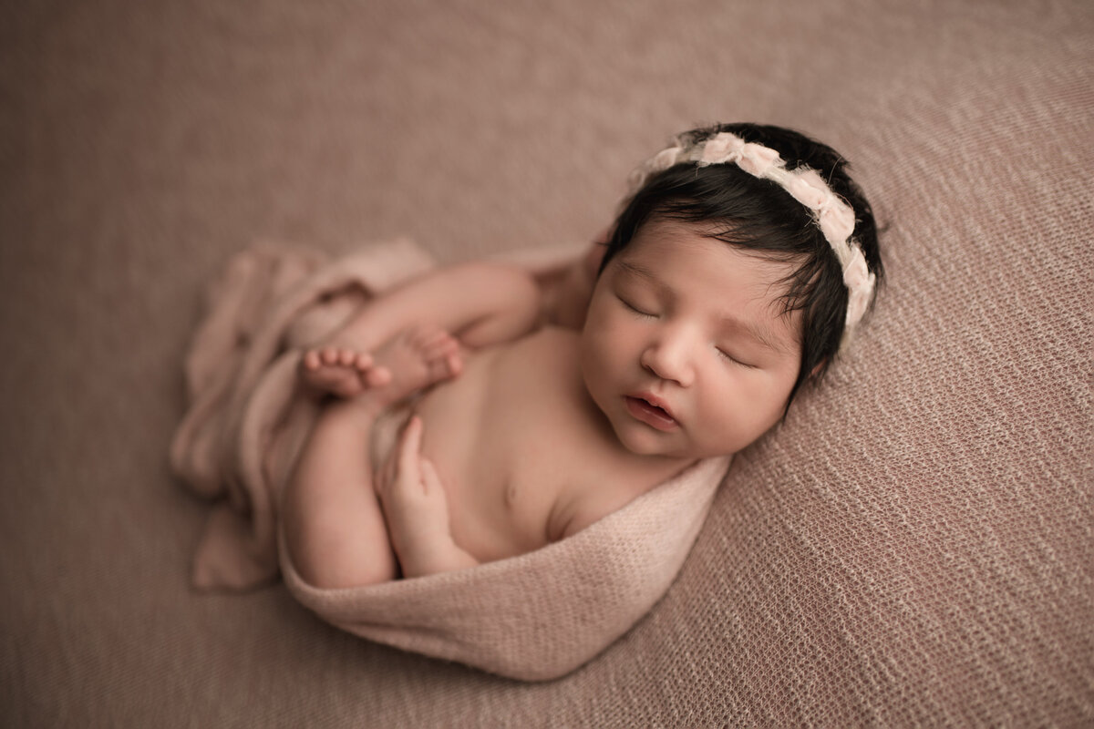 Angled aerial image of Riverside, CA newborn photoshoot. Baby girl sleeping on her back and wearing a pale blush headband. Pale blush fabric is around her body so her legs are folded atop of he. Her hands are resting on her belly.  Captured by Best Riverside, CA newborn photographer Bonny Lynn Photography.