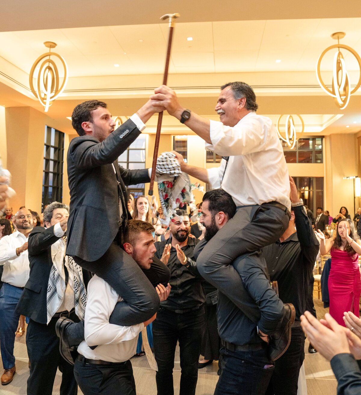 Two men are carried on the shoulders of other men, holding onto a stick. They are facing each other in a large room filled with people who are clapping and cheering. The atmosphere is festive, with warm lighting and elegant decor.