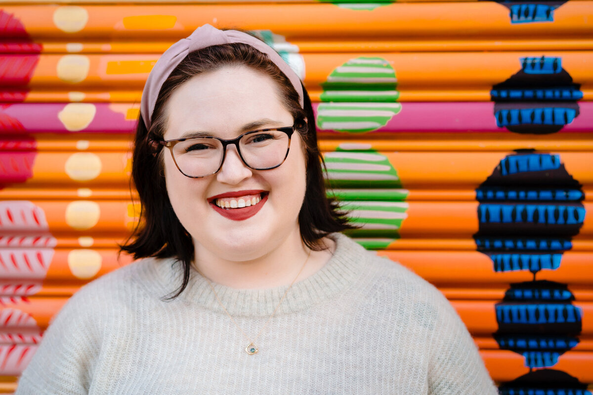 A person in glasses smiling while standing in front of a colorfully painted wall.