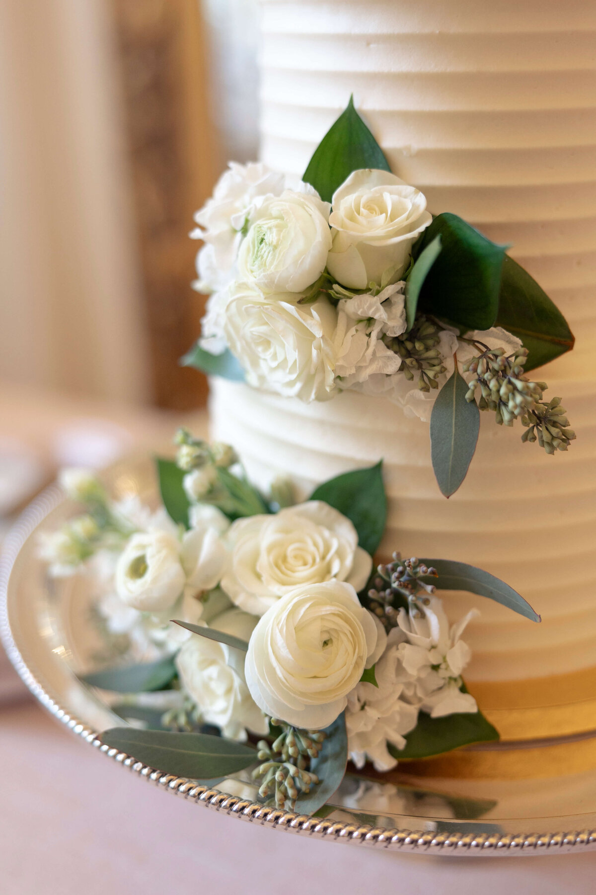 A close-up of a white tiered wedding cake adorned with white roses and greenery, placed on a silver platter with a beaded edge. The cake has a textured, ribbed design on the sides.