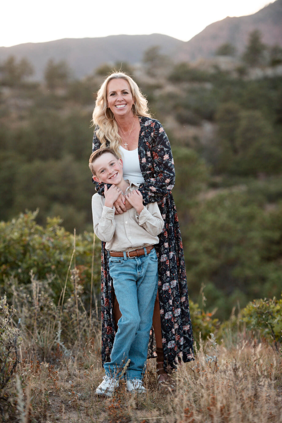 A mother in a floral print dress hugs her son while they stand in a mountain trail