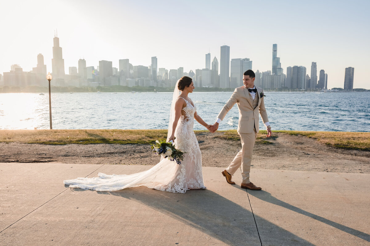 Groom wearing tan suit and his bride walk at sunset with lake Michigan an Chicago skyline in background