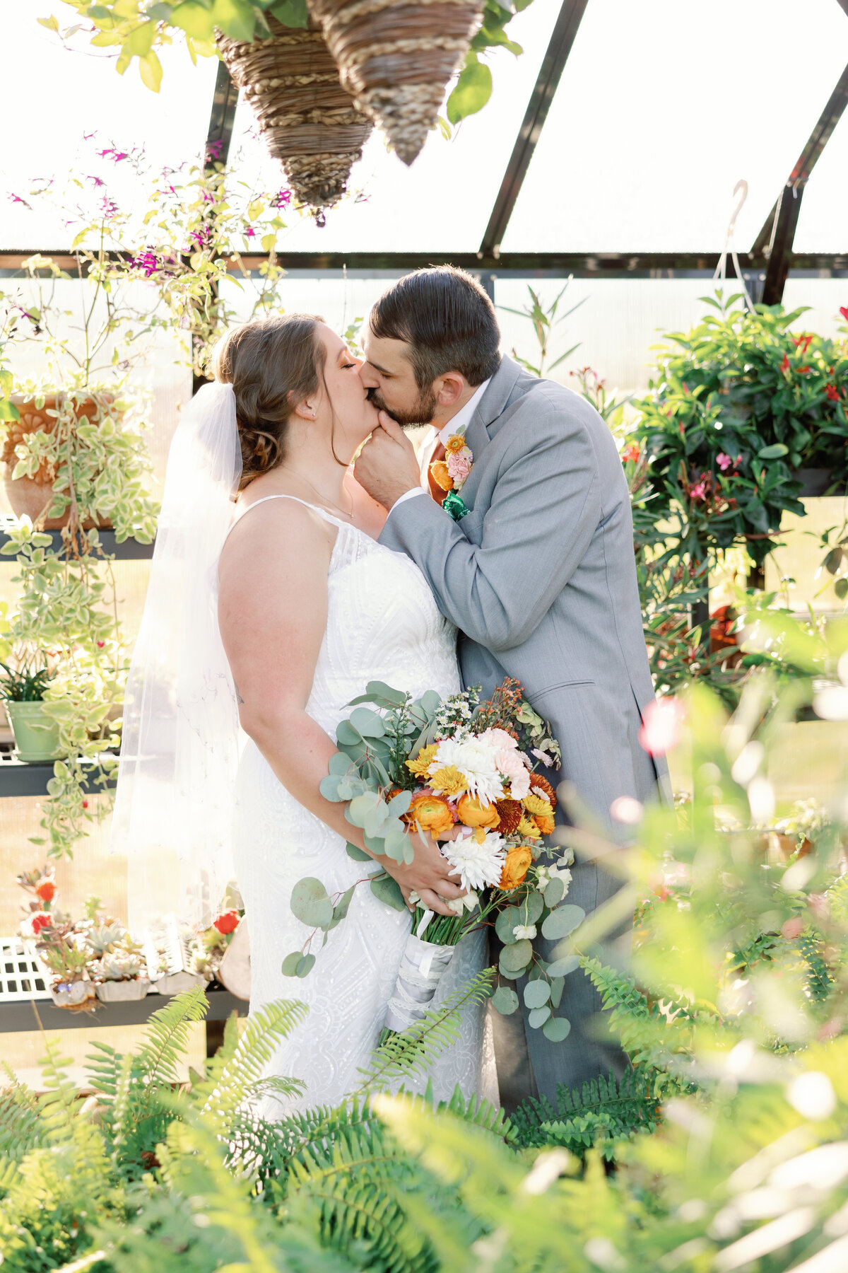 Bride and groom kissing in plant-filled greenhouse with ferns and hanging baskets at Carolina Home and Garden wedding venue