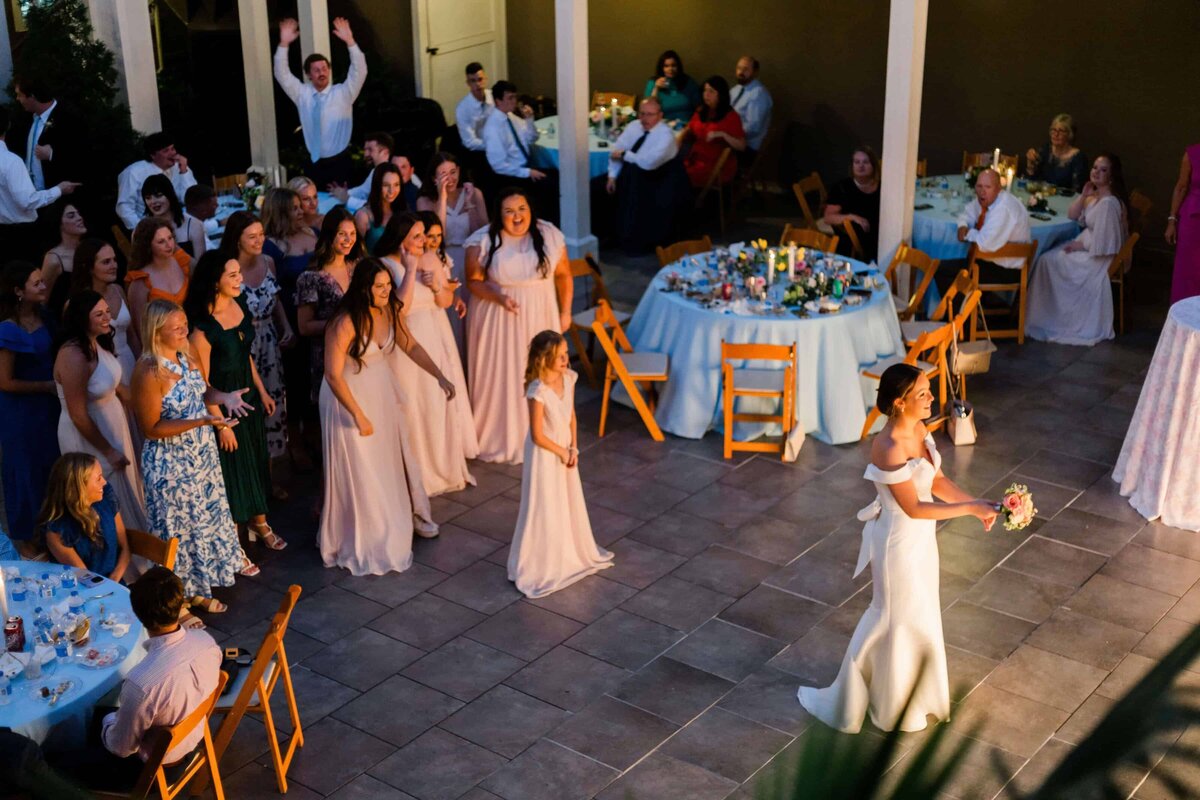 A bride in a white dress prepares to throw her bouquet to a group of excited women and girls gathered behind her at a courtyard wedding reception, with guests seated at decorated tables nearby.