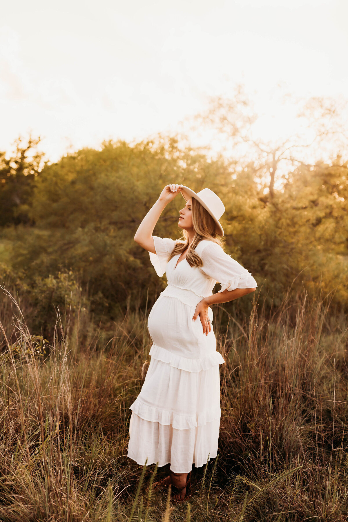 Expecting mother standing to the side in a white dress with one hand on her back and one hand holding her hat