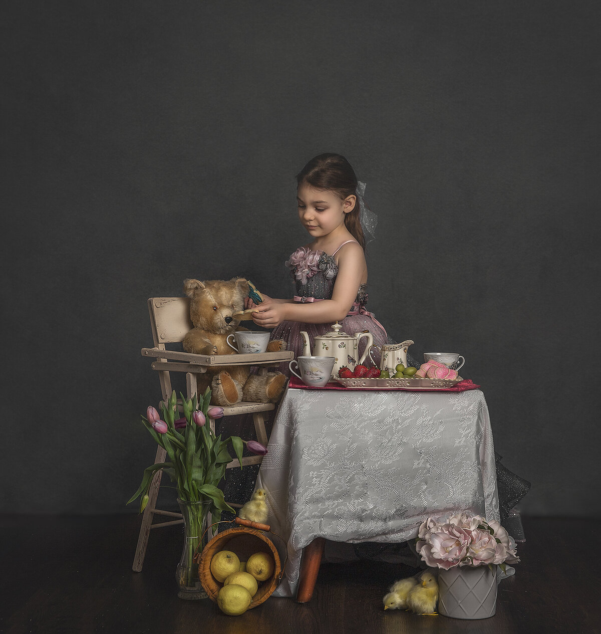 A young girl in a pink and gray gown feeds her teddy bear at a picnic.