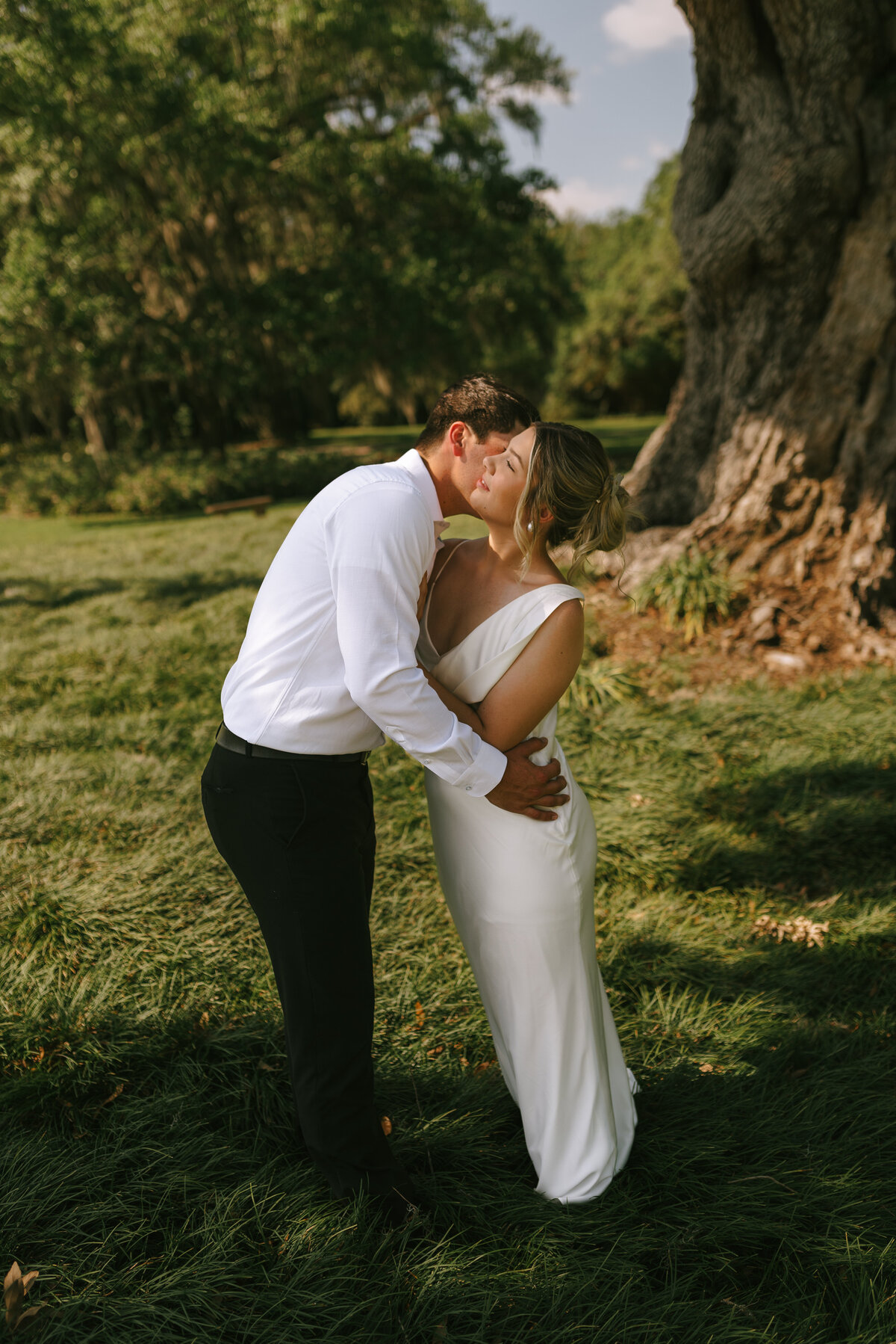 a true to color photo of a wedding couple leaning in close to each other in a grassy area under the moss draped oak trees of louisiana.