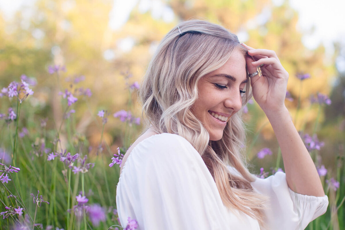 A woman with long blonde hair smiles while standing in a field of purple flowers. She is wearing a white top and has one hand gently touching her forehead. The background is blurred, showcasing greenery and soft sunlight.