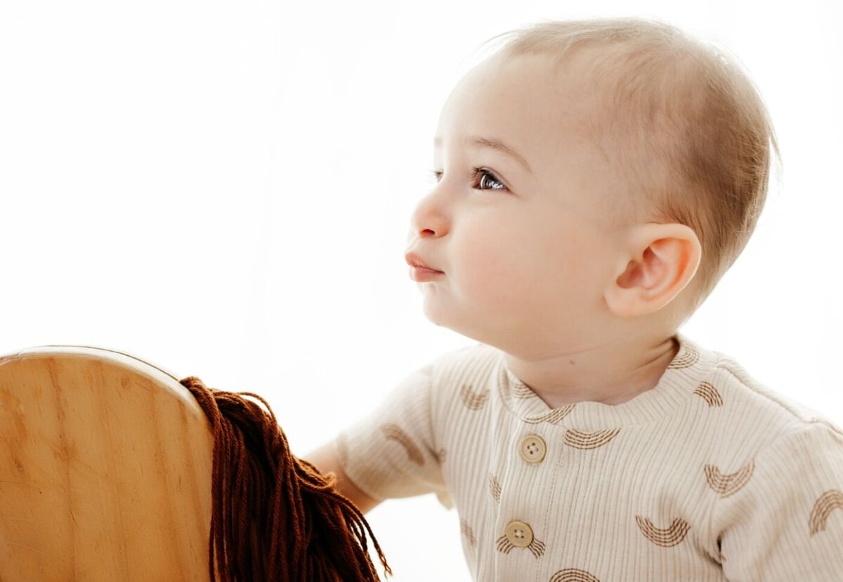  a baby with fair skin and light hair, looking off to the side with a thoughtful expression near a wooden rocking horse.