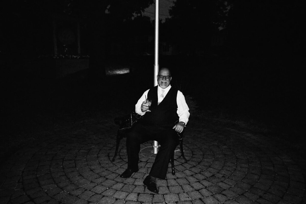 A man in a suit and vest sits alone on a metal chair in the center of a circular stone patio at night, holding a drink and smiling. Captured by an NJ wedding photographer, he’s illuminated in the darkness, creating a cinematic moment.