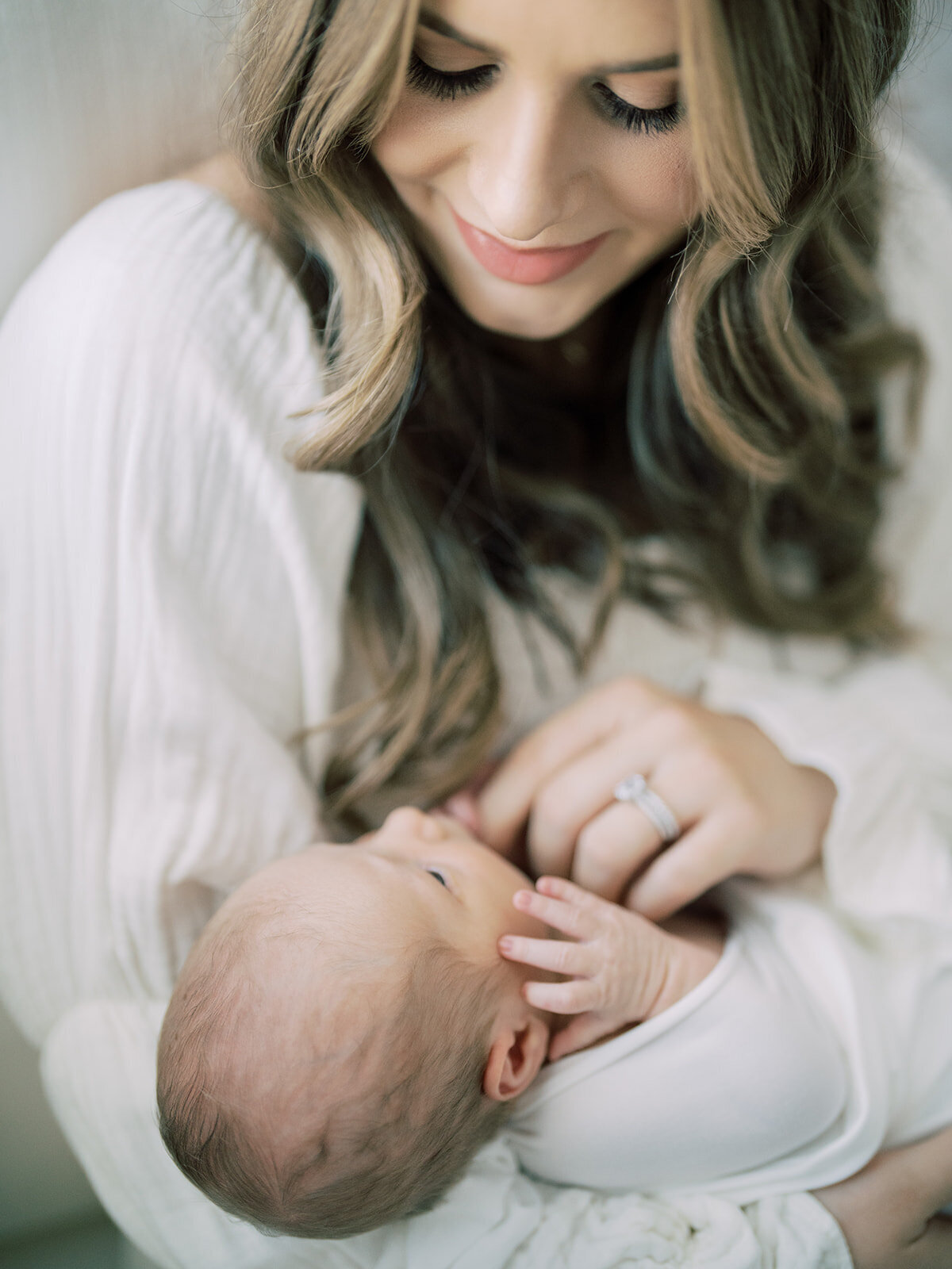 Mother with brown hair and white dress holds her newborn baby who is swaddled in white and smiles down at her.