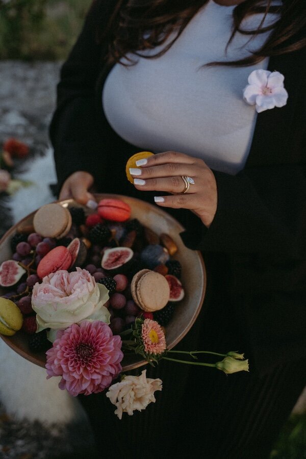 Anissa piochant un macaran dans une corbeille remplie de fruits, de fleurs. Instant gourmand immortalisé par Kamescop, photographe séance d'engagement.