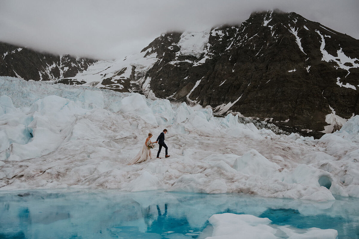 Couple walking hand in hand above an aqua blue glacial tide pool on the Knik Glacier .