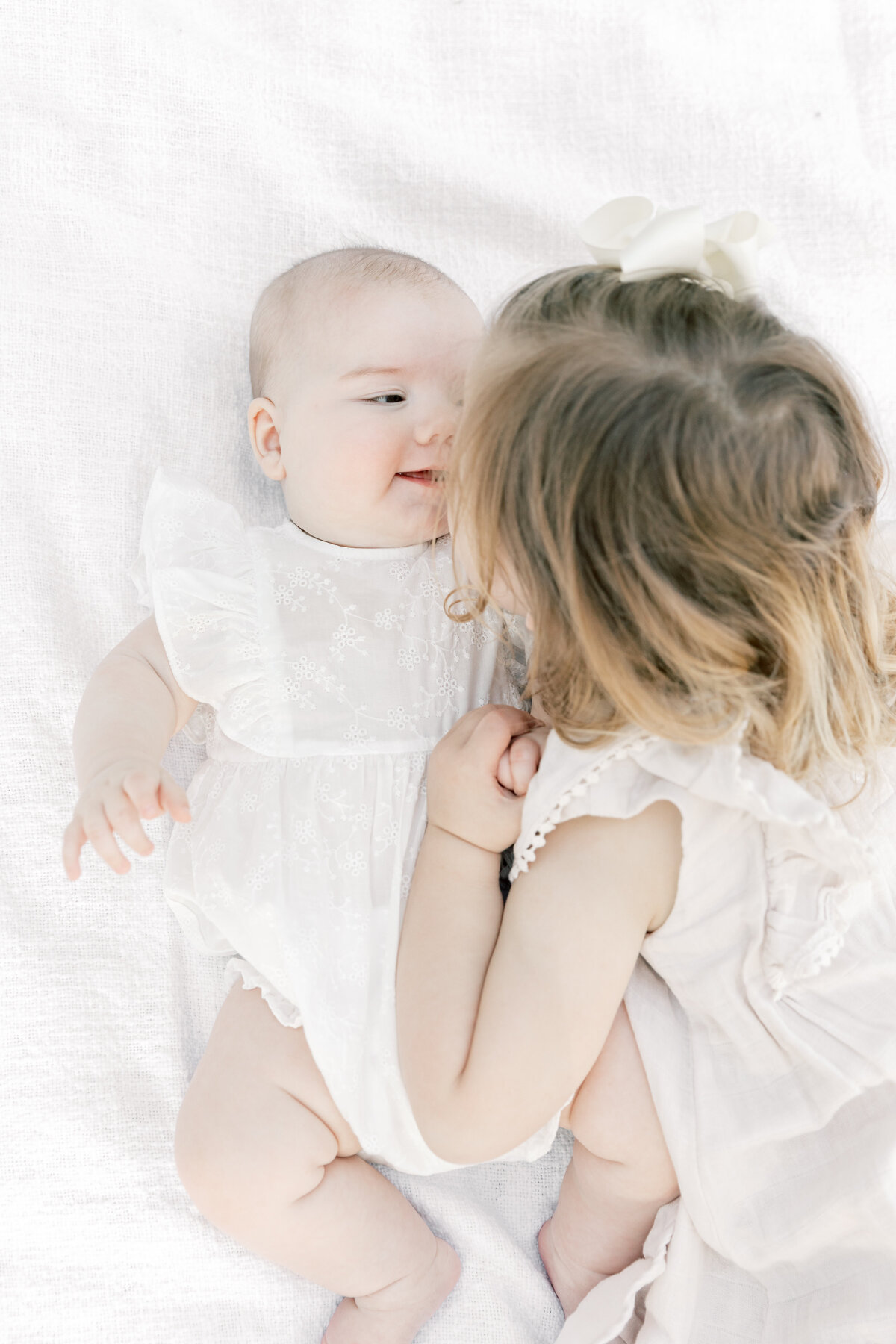 A toddler girl in a white dress leans in for a kiss to her newborn baby sister taken by a Charlotte Family Photographer
