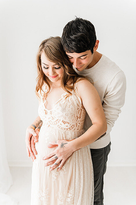 Couple shares a quiet moment while holding her baby bump, dressed in light tones with soft natural light pouring in.