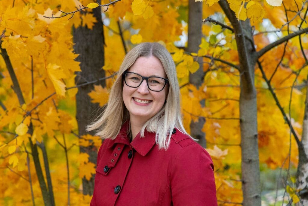 Jenny Moran standing near a rustic wooden fence, surrounded by golden autumn leaves.
