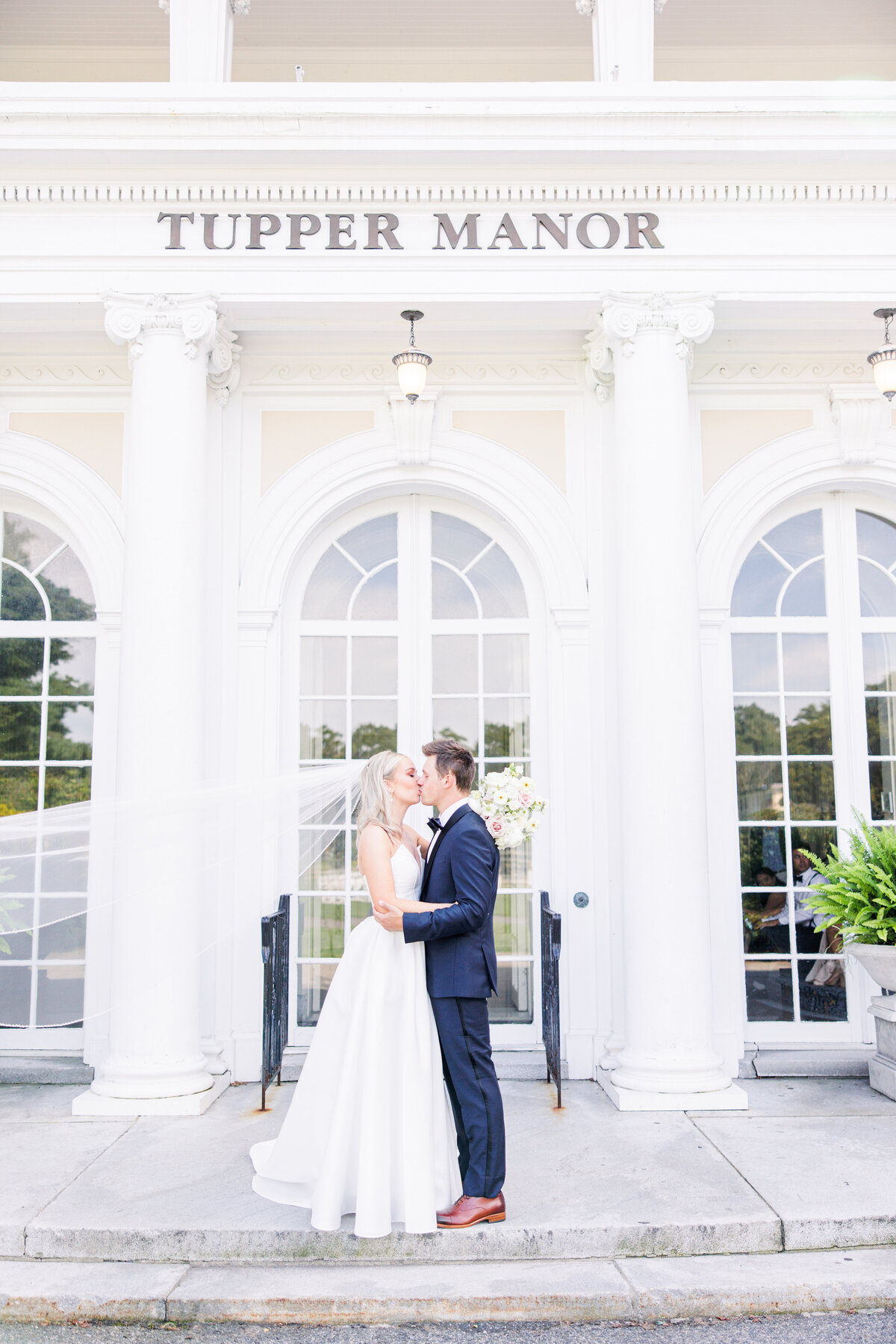 Bride and groom kissing with veil blowing in front of Tupper Manor