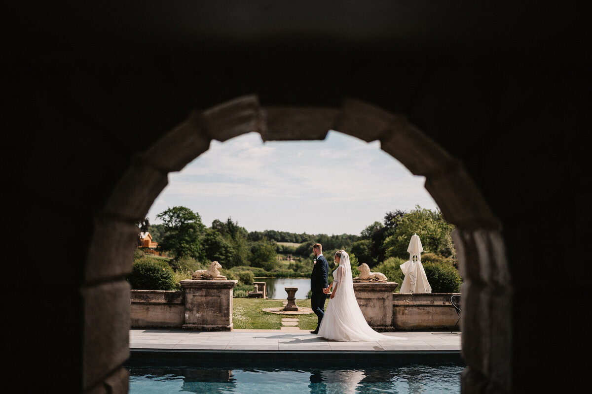 Bride and groom holding hands by the poolside at Foxtail Barns, Stoke, framed through a stone archway with countryside views and elegant wedding details.