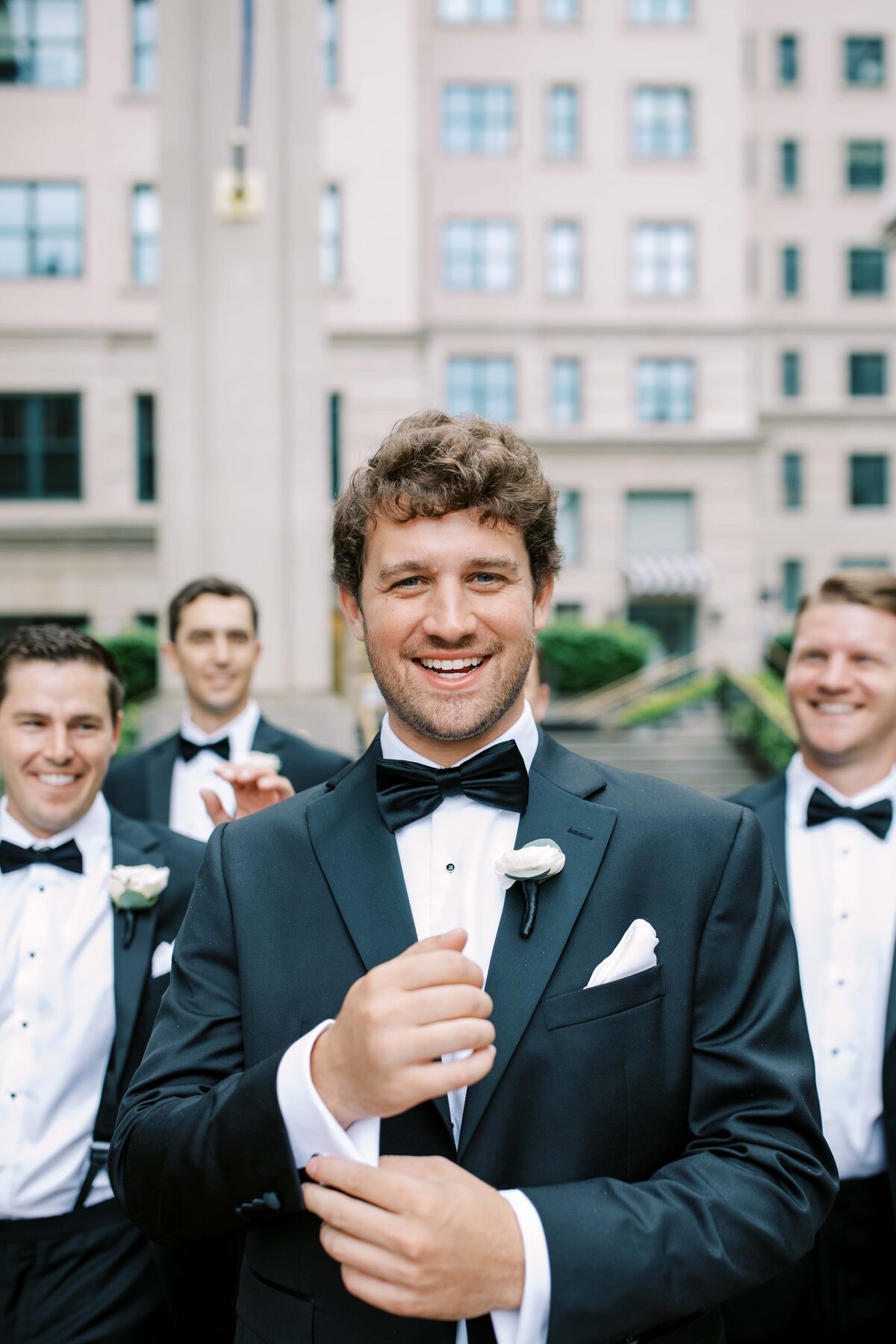 Handsome white groom in a black tux smiling directly at the camera while fixing his cufflink with the groomsmen walking behind him outside of The Willard Hotel in washington DC