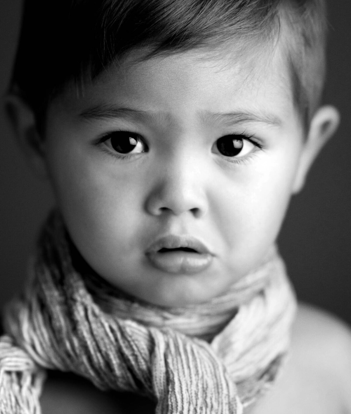 Black and white portrait of a young child with a curious expression. The child has short hair and wears a textured scarf, conveying a soft, contemplative mood.