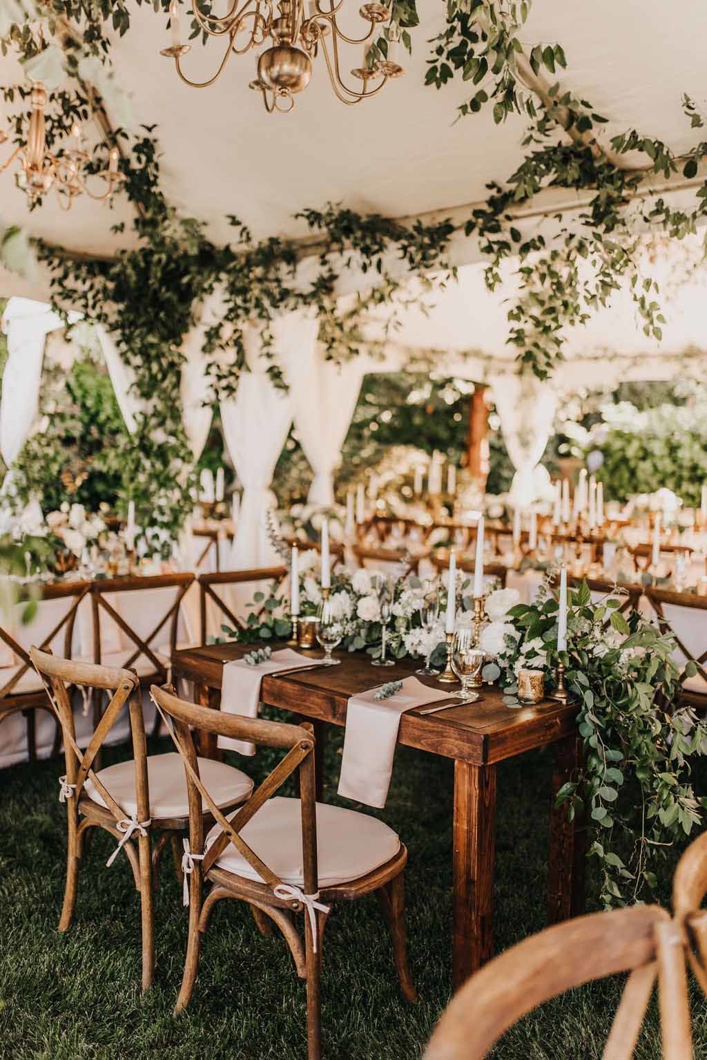 wooden sweet heart table covered in greenery in wedding tent with smilax greenery in ceiling