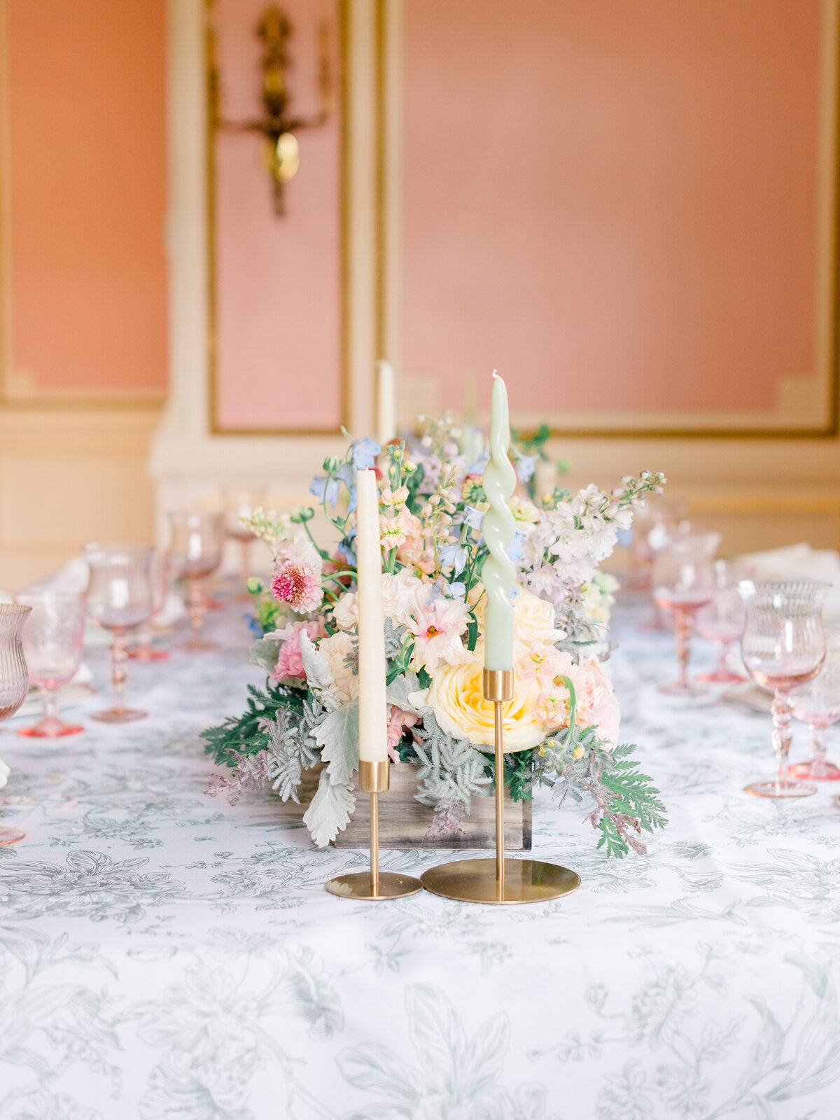 Close-up of pastel wedding centerpiece with blush, yellow, and ivory florals on a toile-covered reception table in the pink room at Cairnwood Estate. Twisted taper candles in gold holders and blush pink glassware add whimsical elegance. Captured by Philadelphia Fine Art Wedding Photographer Elizabeth Kane for a luxury spring wedding editorial in Bryn Athyn, Pennsylvania.