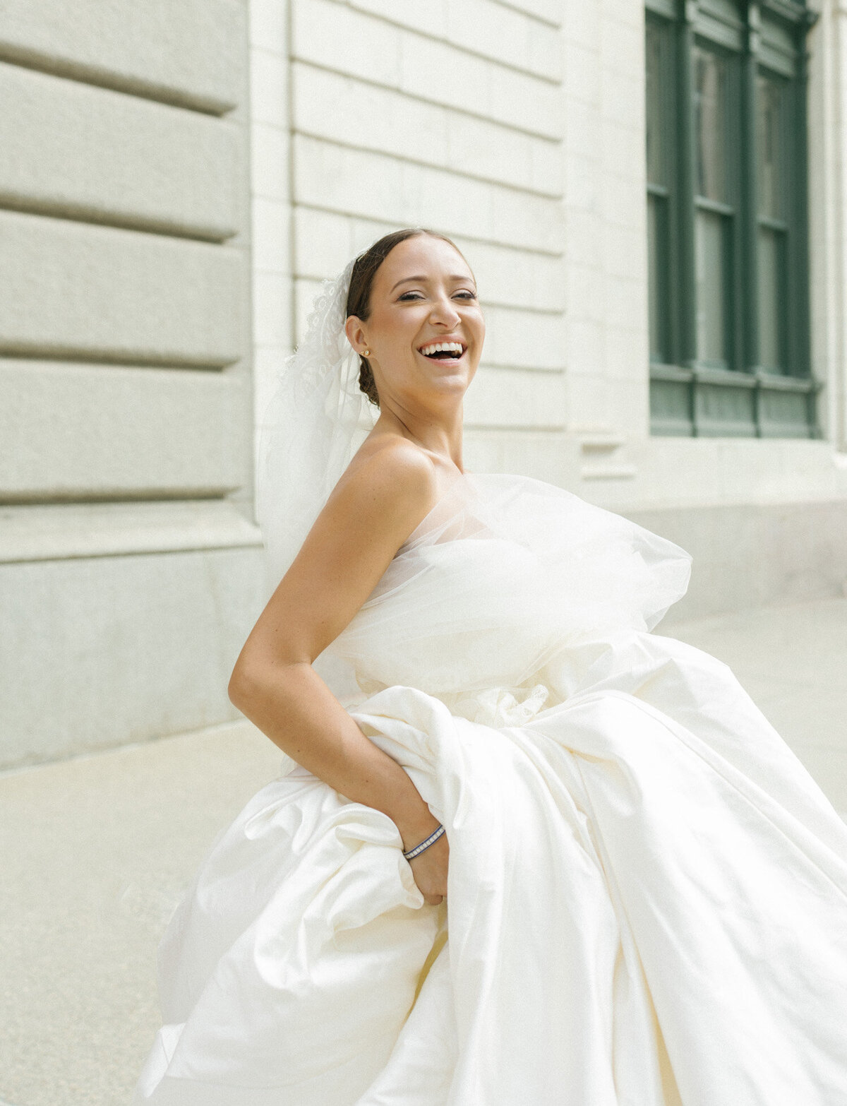 a bride in a long cathedral veil carrying her silk ballgown laughs in Detroit, Michigan