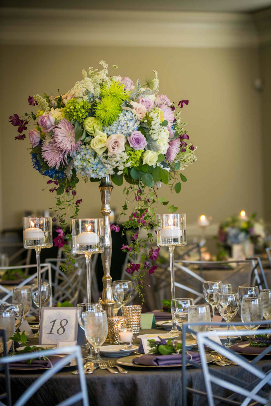 large centerpiece of blue hydrangea, purple roses, green chrysanthemums, , and trailing purple clematis, with floating glass candle holders