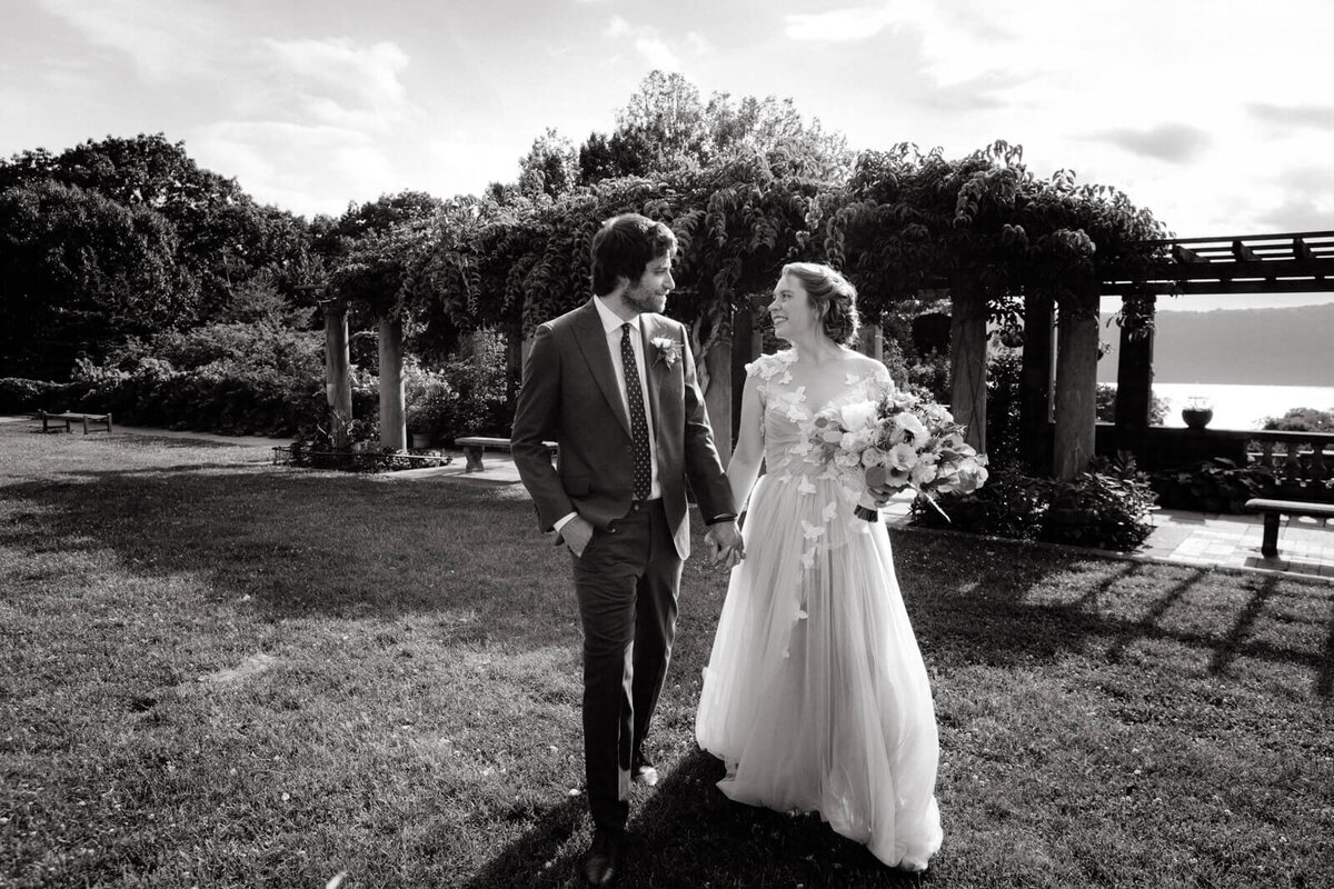Black and white photo of the bride and groom, walking while holding hands and looking at each other sideways, at a garden.