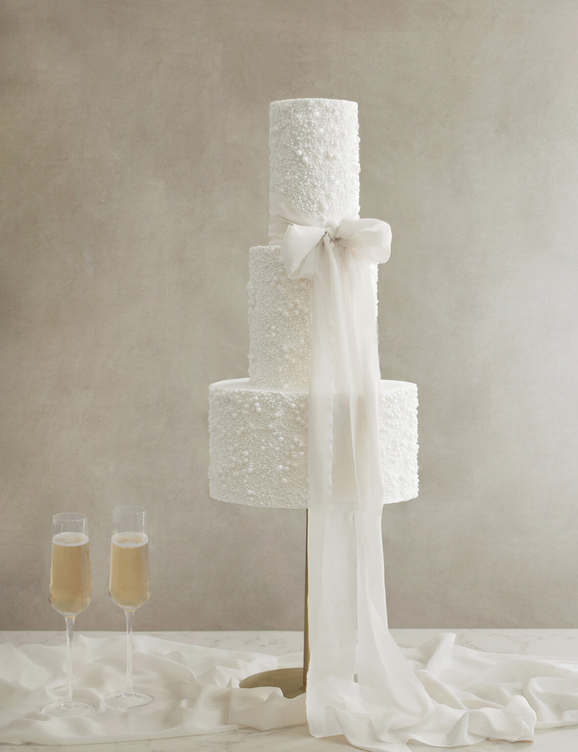 A white three tiered cake with a hand died white silk bow with two glasses of champagne