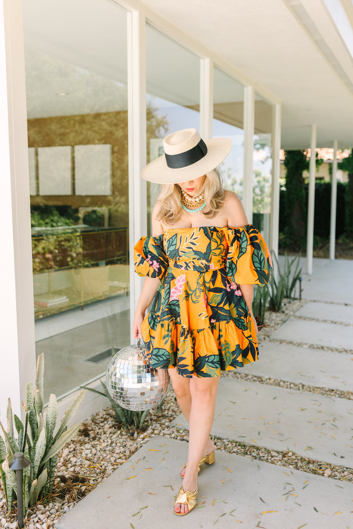 Vibrant fashion-forward branding image of a woman in a yellow floral dress and wide-brim hat holding a disco ball outside a midcentury modern home — playful and stylish.