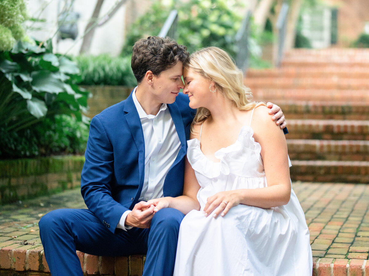A man in a blue suit and a woman in a white dress sit close together on brick steps, surrounded by greenery. They are gazing into each others eyes with gentle smiles, exuding a sense of romance and connection.