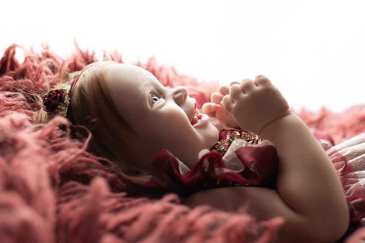 A smiling baby girl in a burgundy and gold outfit lies on a fluffy red blanket, captured in warm light during a Dallas milestone photography session.