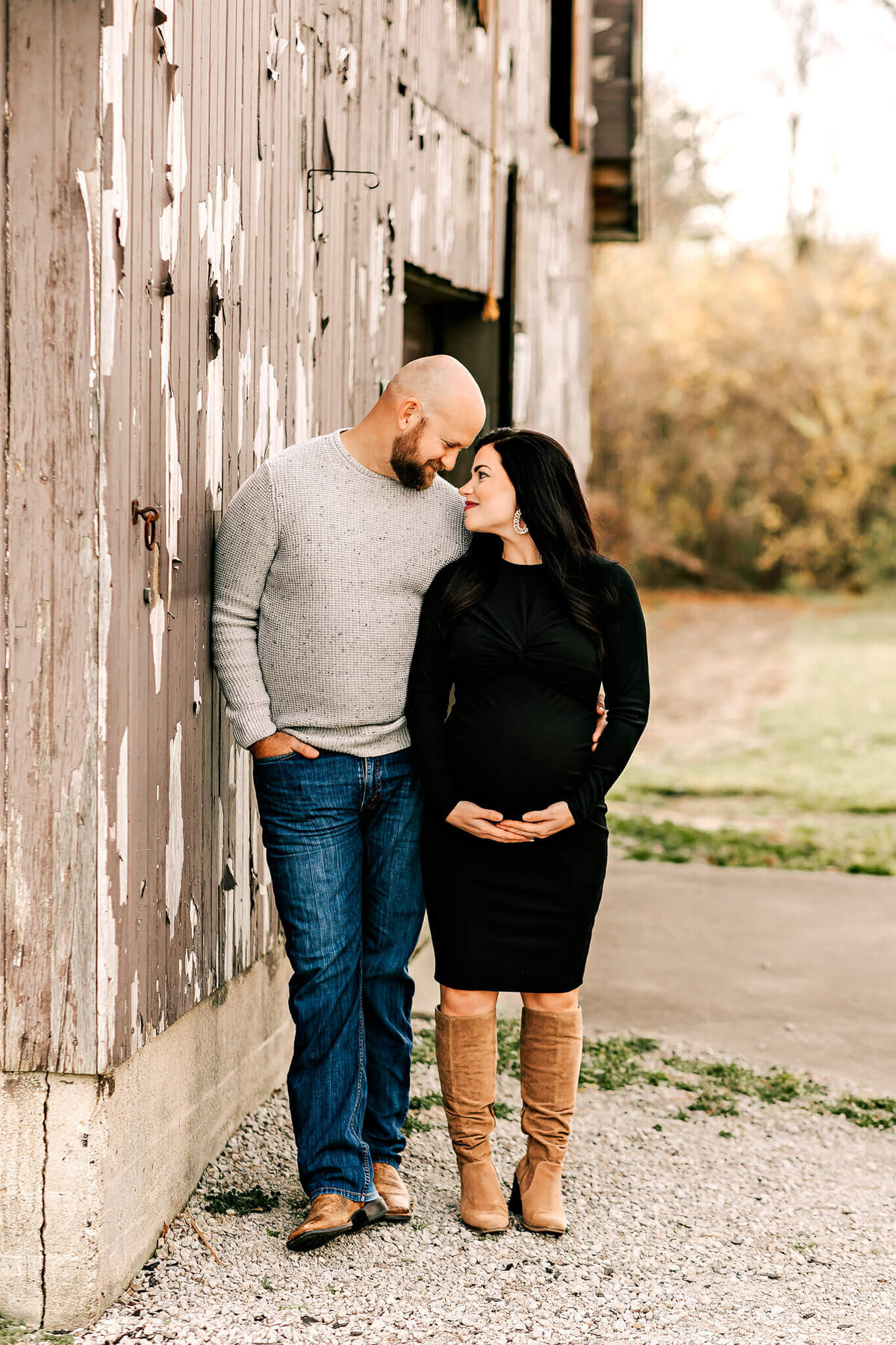 A couple stands closely by a weathered wooden wall, gazing lovingly at each other in this intimate example of maternity portraits. The pregnant woman cradles her belly as the man in a gray sweater smiles warmly—captured by a Bloomington maternity photographer.