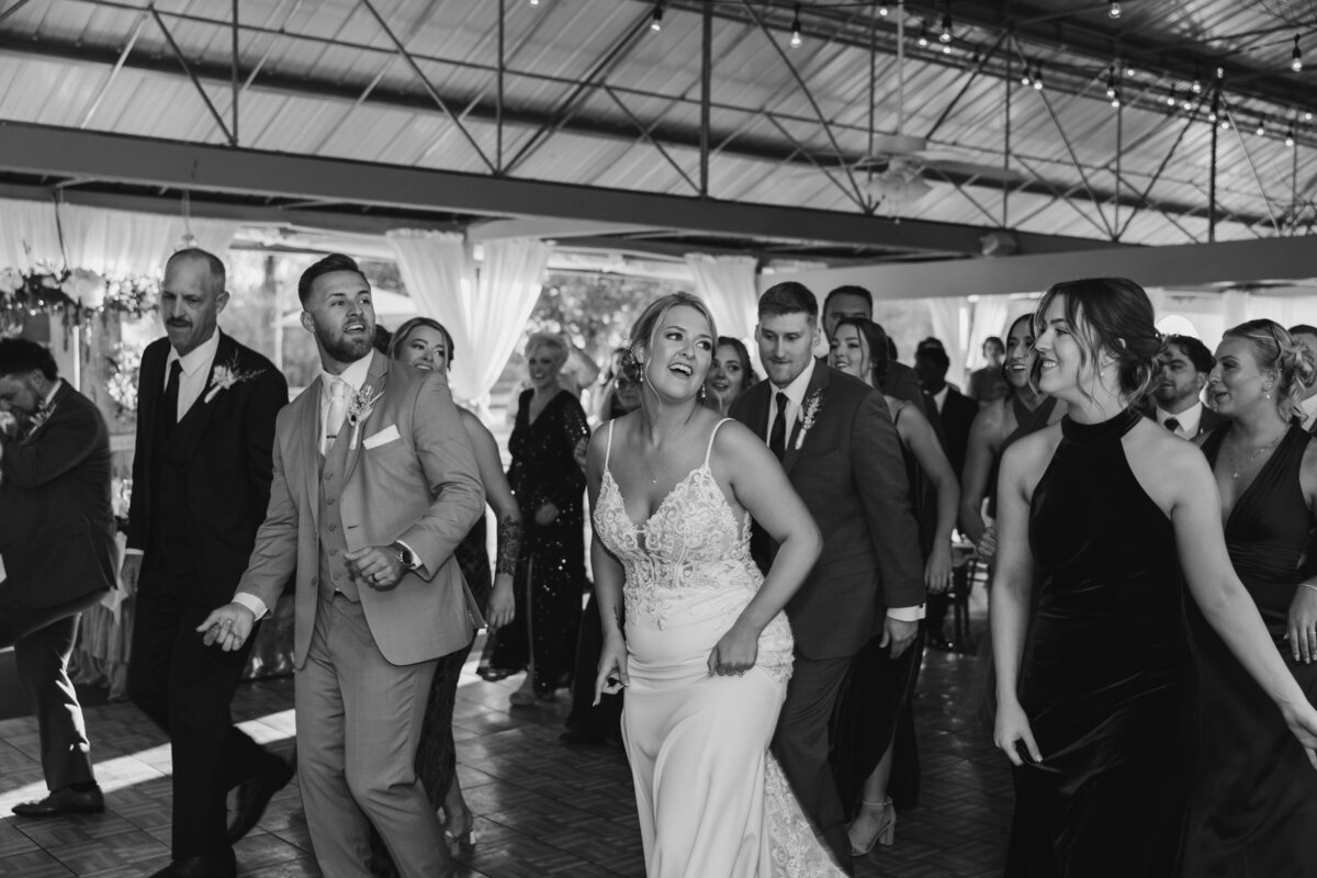 A black and white photo of a lively wedding reception at Lippincott Manor. The bride, wearing a lace gown, dances joyfully alongside the groom in a light-colored suit, surrounded by their wedding guests. The group is engaged in a coordinated line dance, with smiles and laughter filling the room. The background features draped curtains and elegant lighting, adding to the celebratory atmosphere.