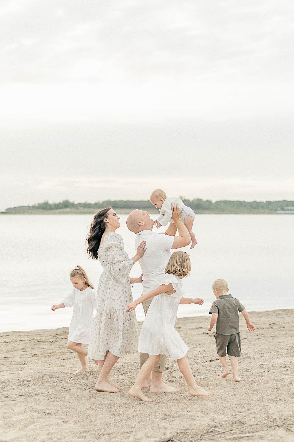 Family of six plays barefoot along the shoreline of a Kansas City lake, all dressed in light neutral tones during their baby's sitter milestone session.