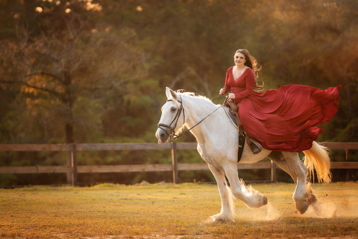 Rodeo queen on a white shire in the outdoor arena.  There is a wooden fence behind them.  The horse is running.  The woman riding him is wearing a long red dress that is blowing in the wind.  The horse is running and kicking up dust.