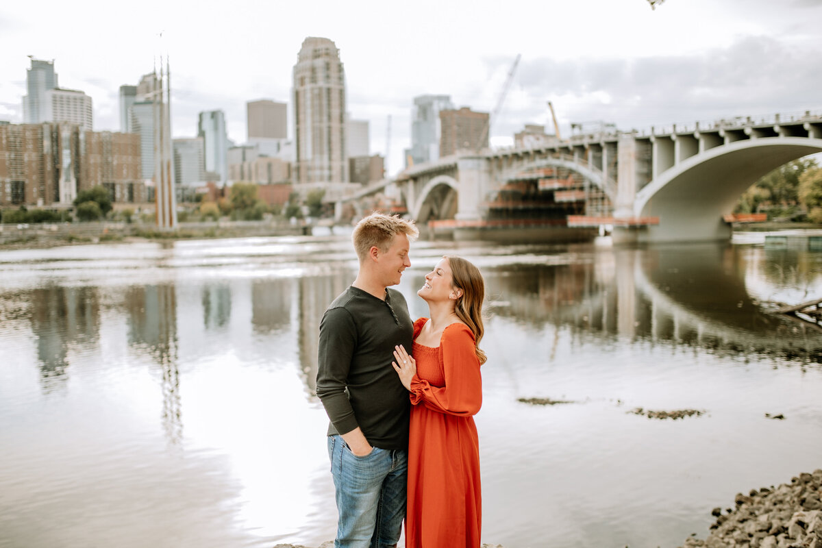Minneapolis St. Paul engagement and wedding photographer, Emmie Verdell Photo, documents a couple lovingly gazing at one another with Stone Arch Bridge and a skyline in the background.