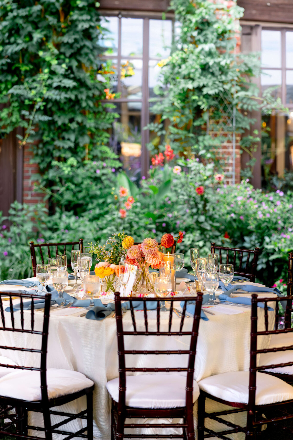 An elegantly set round table with white tablecloth and dark chairs is adorned with colorful flowers and candles. The background features lush greenery and flowers climbing a brick wall, with large windows reflecting the outdoor scene.