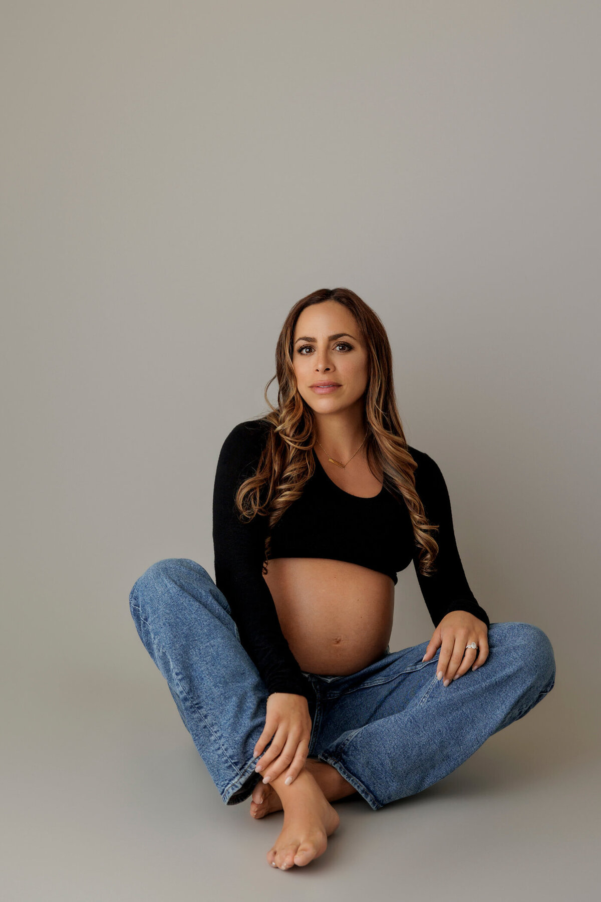 A pregnant woman sits on the floor against a neutral gray backdrop, wearing a black long-sleeve crop top and loose-fitting blue jeans that reveal her baby bump. She sits cross-legged with one hand resting on her knee and the other gently touching her belly, gazing directly at the camera with a calm and confident expression. The casual and relaxed pose highlights the natural beauty of her pregnancy.