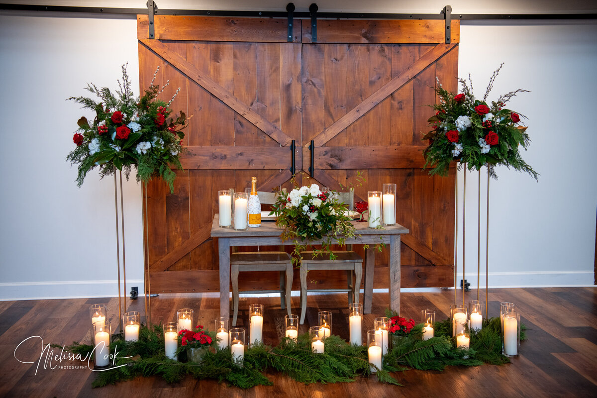A rustic wedding setup features a wooden table with flowers and candles. Red and white floral arrangements adorn the scene, and a wooden barn door serves as the backdrop. Candles line the area, creating a warm, romantic ambiance at this charming Leesburg VA wedding venue.