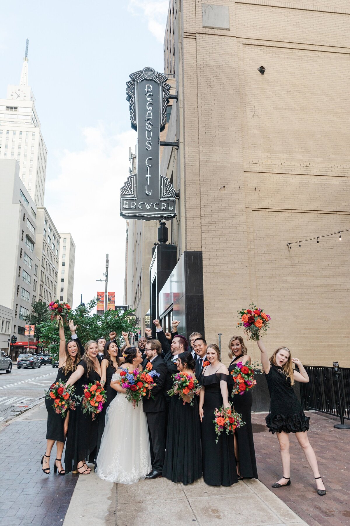 A portrait of a bride and groom looking joyfully at each other while backed by their wedding party who are cheering them on in celebration after their wedding ceremony at the Pegasus City Brewery in Dallas, Texas. The bride is on the left and is wearing a long, intricate, white dress and is holding a large bouquet. The groom is on the right and is wearing a black suit with a tie and boutonniere. The wedding party is wearing either black suits or black dresses with bouquets.