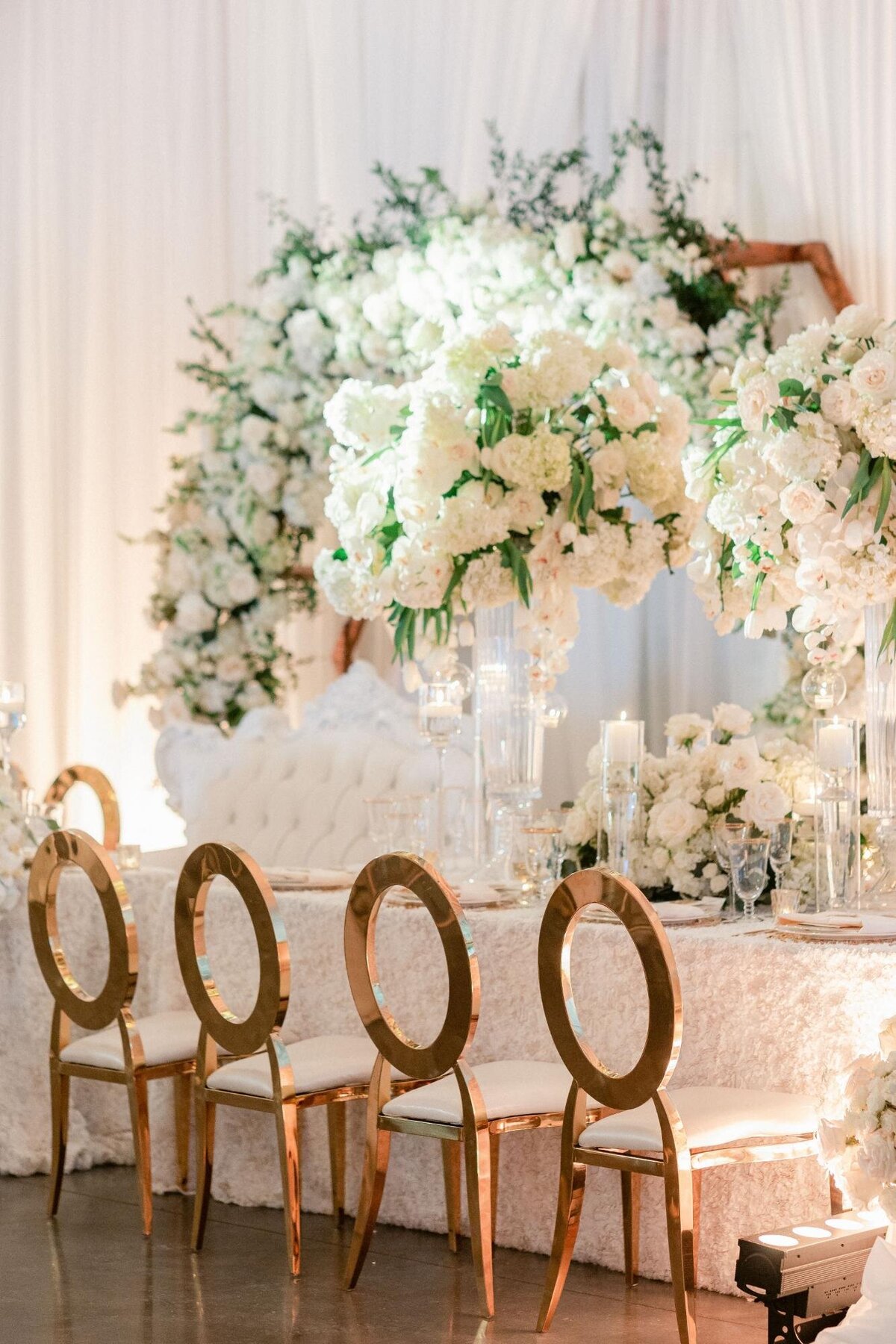 Elegant wedding reception setup with white floral arrangements and gold-accented chairs.