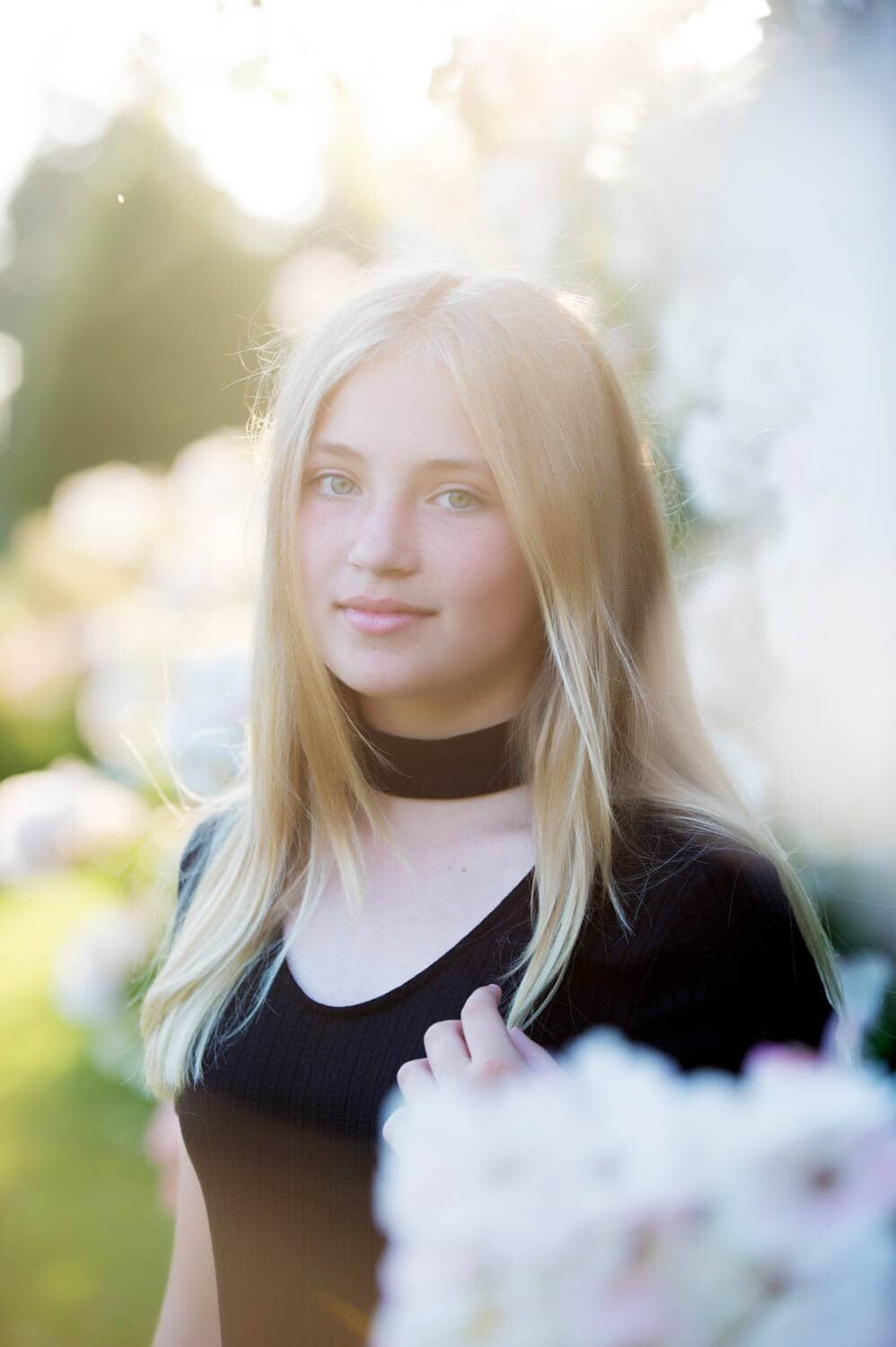 professional headshot of Young woman with blonde hair in a black shirt, standing near white flowers.