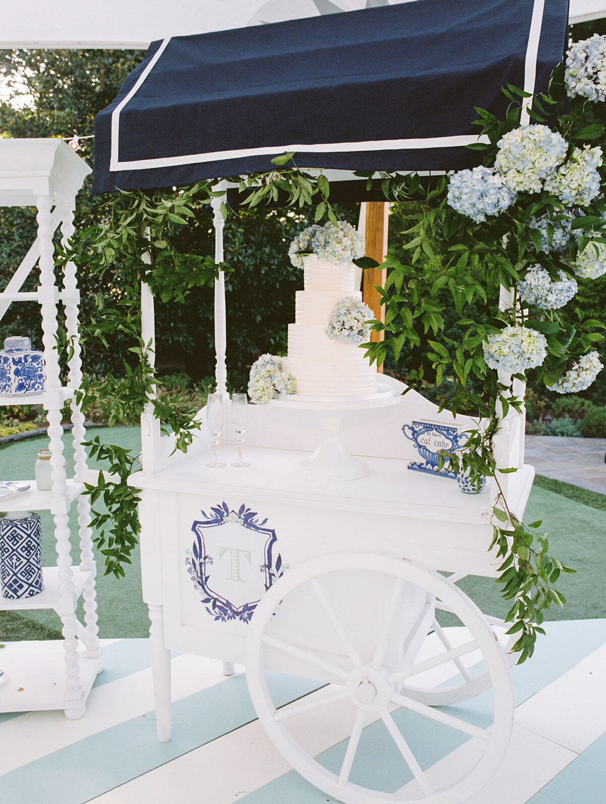 The photo was taken at The Hamptons, New York, waterfront estate tented wedding by photographer Bonnie Sen. In the picturesque setting, a white decorative cart catches the eye with its blue and black canopy, embellished with fresh green foliage and clusters of beautiful hydrangea flowers. Noteworthy is the monogrammed T in a charming blue floral design adorning the cart. On top of the cart, an elegant tiered white cake takes center stage alongside a delicate blue teacup, adding a touch of sophistication to the refined setup. East West Films Videographers were also present to capture the special moments of this beautiful wedding.