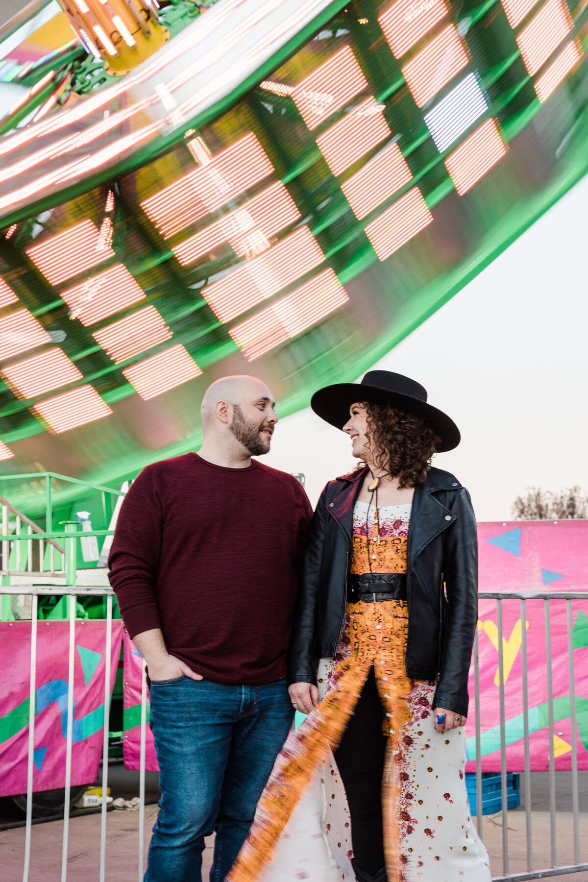 A couple holding hands and looking into each other's eyes in front of a rapidly spinning carnival ride during their engagement shoot at a carnival in DFW, Texas. The woman on the right is wearing a large brimmed black hat, black leather jacket, and an intricately detailed and colorful dress. The man on the left is wearing a maroon sweater and jeans.