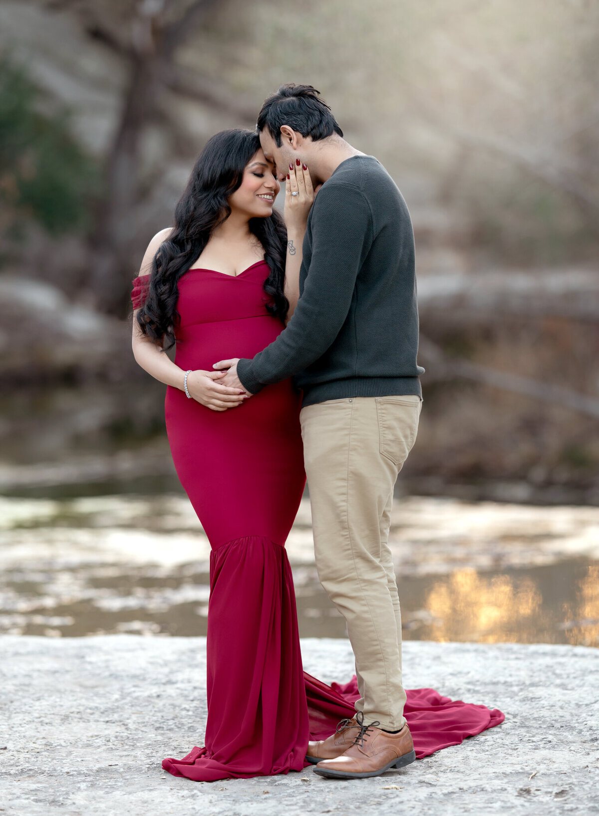 Romantic couple maternity photo in Austin with the mother-to-be in a fitted red gown, sharing a tender moment with her partner by a serene creek.