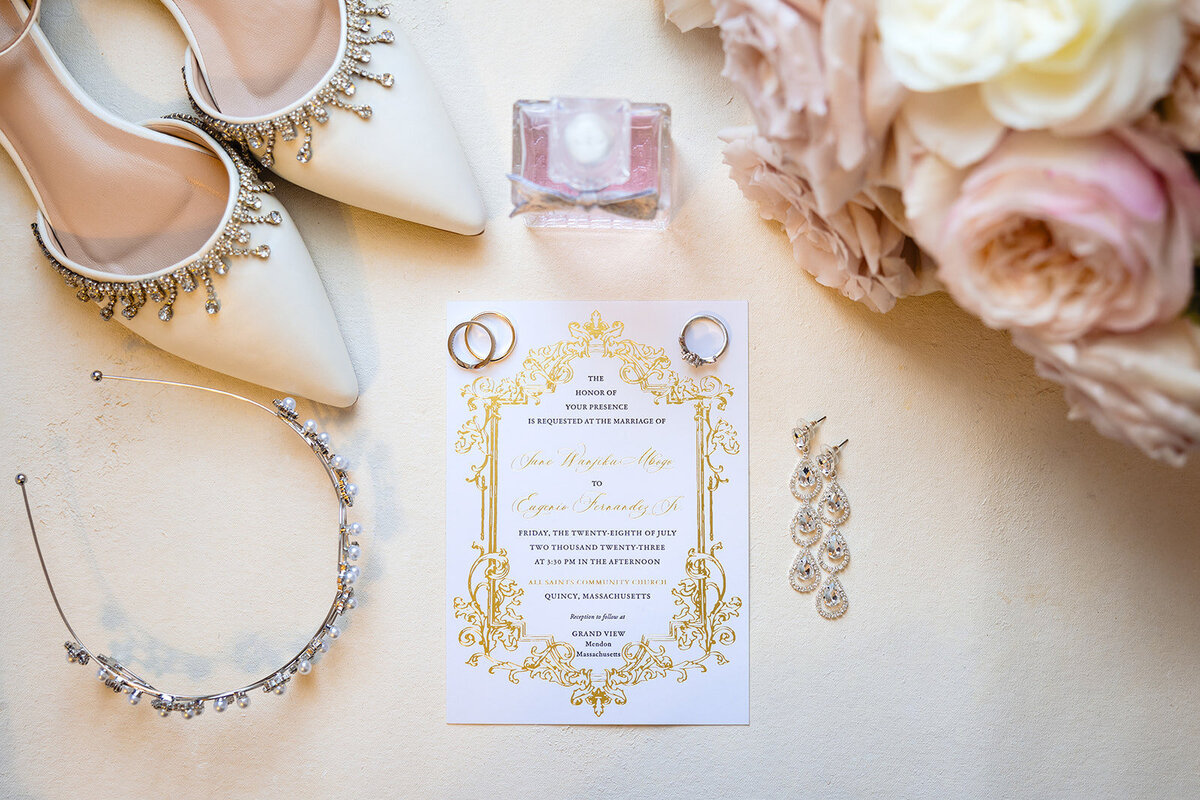 A flat lay of wedding accessories with elegant ivory shoes adorned with rhinestones, a bottle of perfume, wedding rings, a pair of earrings, and a floral wedding invitation stating a request for the honor of presence at the marriage of 'Alice Hawthorne May' to 'Gregory Li' in Quincy, Massachusetts.