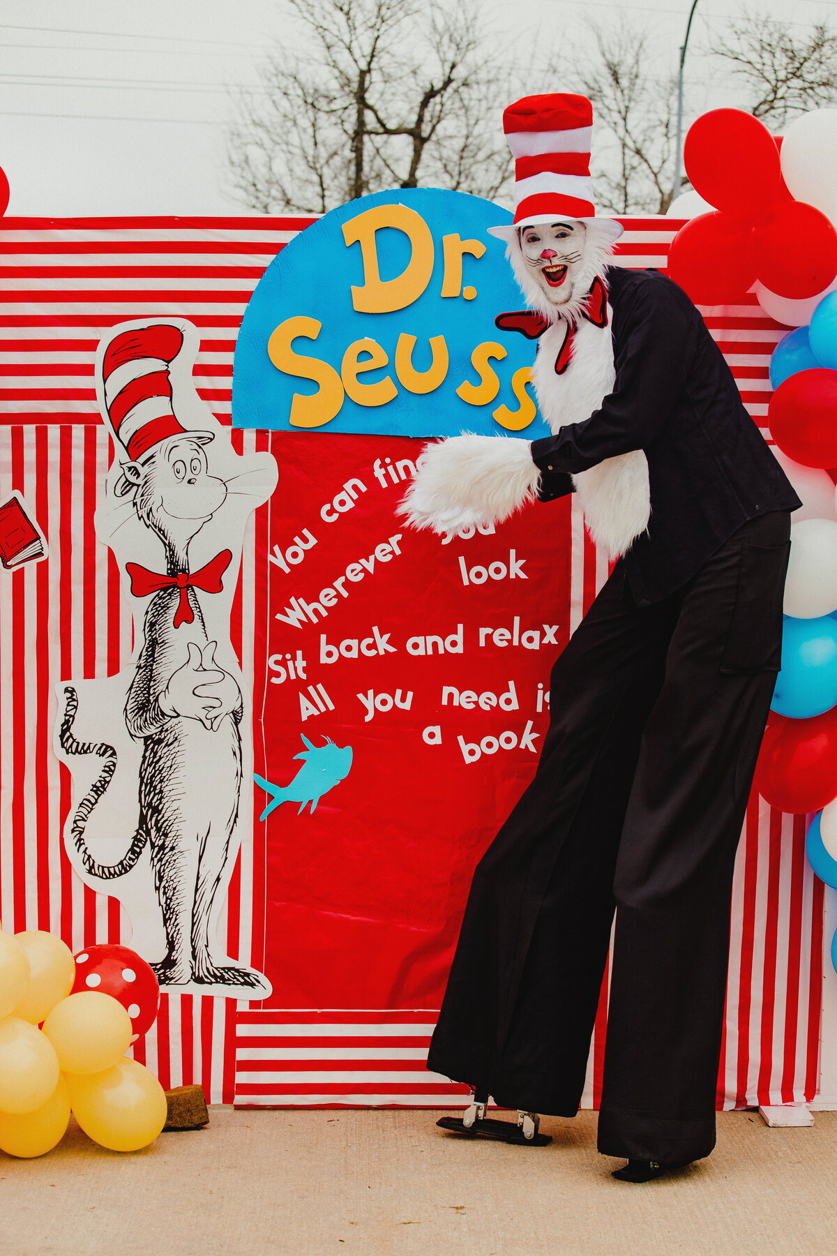 Person wearing a Cat in the Hat costume standing in front of a red, blue and yellow Dr. Seuss themed backdrop display