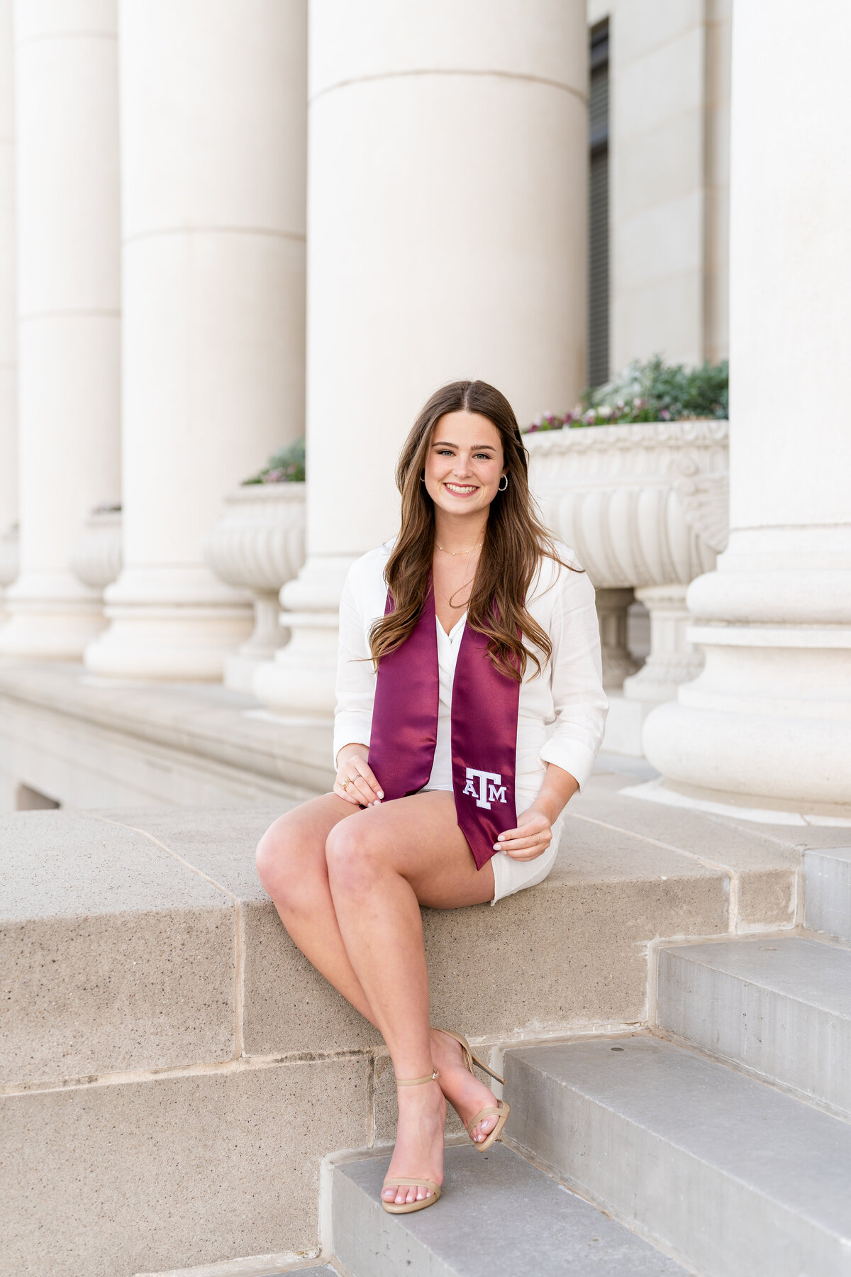 Texas A&M senior girl sitting on Administration Building stairs while smiling and holding stole and wearing white dress